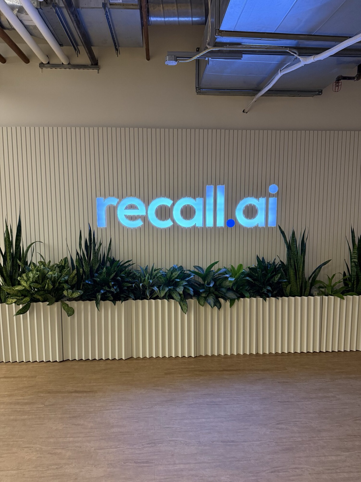 Backlit recall.ai logo on a slatted wall above a row of planters at the office entrance