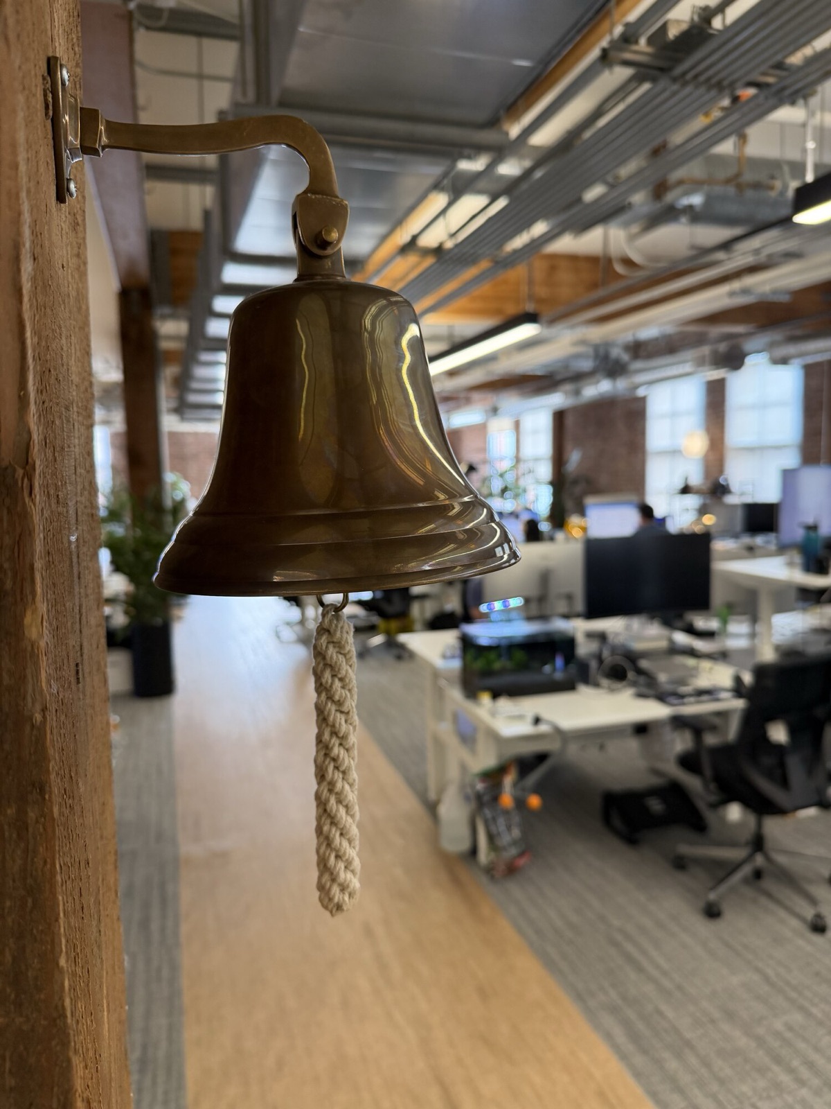 A brass bell mounted on a timber post, with rows of desks and monitors behind it