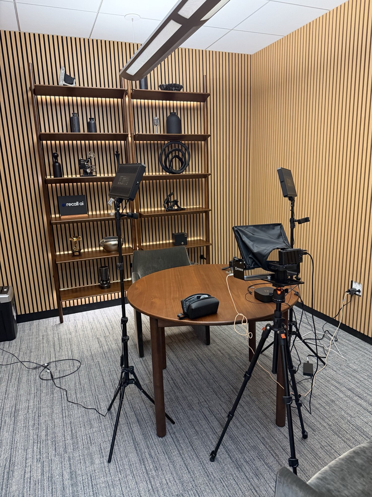 A recording room with a round wooden table, a microphone on a boom stand, a camera on a tripod, and a slatted wood wall