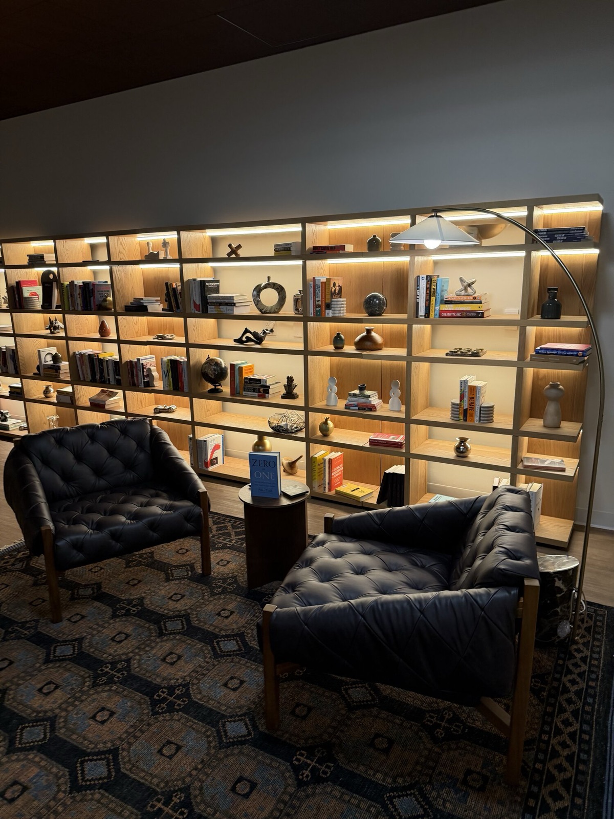 A backlit library wall of shelves with books and objects, two dark leather armchairs in front