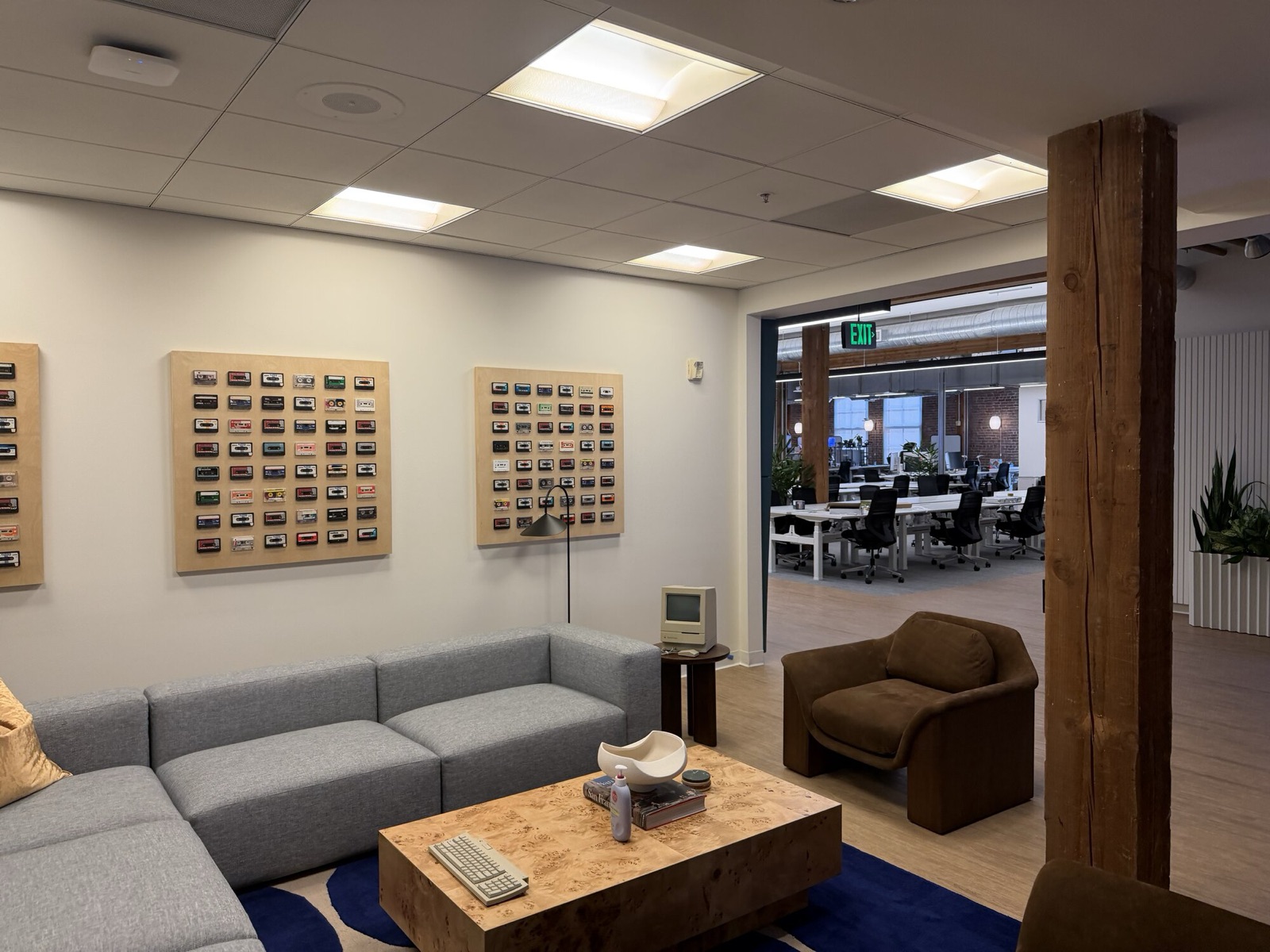 Lounge with grey sofas and a wooden table in the foreground, empty rows of desks and the logo wall behind