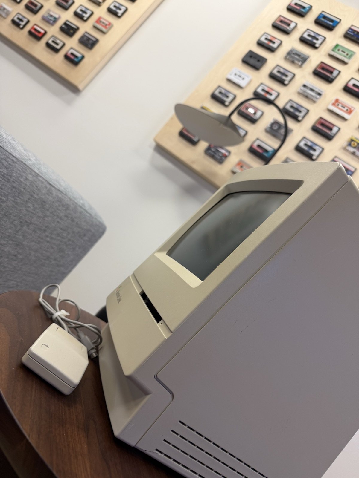 Close-up of a compact Macintosh on a wooden side table, cassette tapes mounted on the wall behind